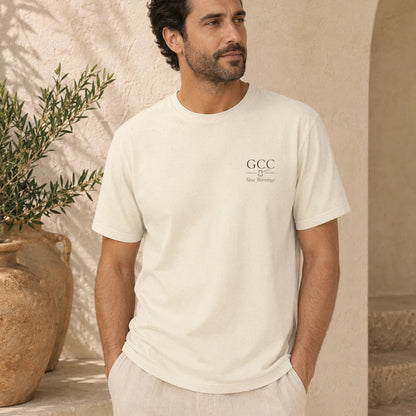 A man wearing a cream-colored short-sleeve t-shirt with a small black 'GCC' logo on the left chest, standing in a warm, sunlit room with beige walls and a potted olive tree.