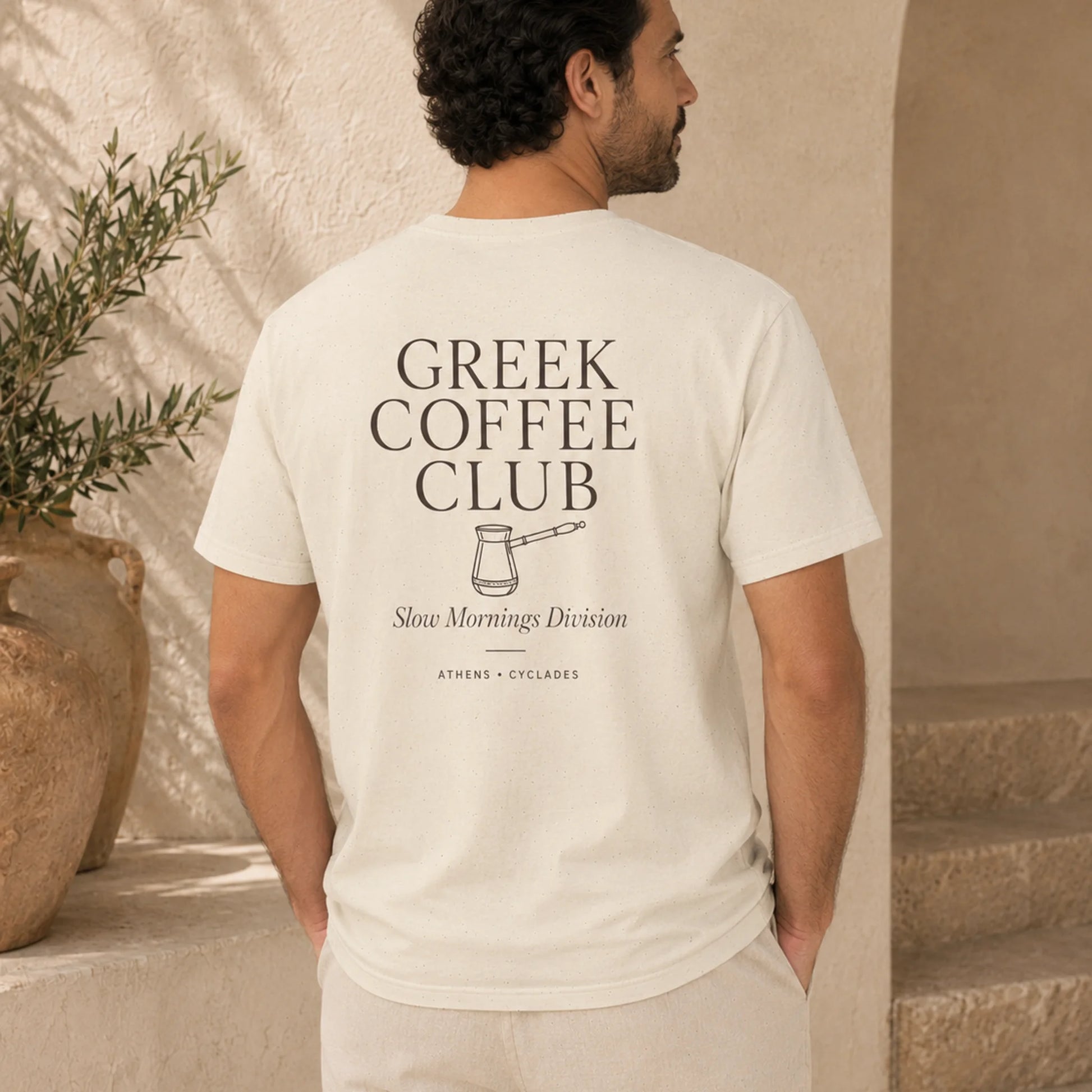 A man viewed from behind wearing a cream-colored t-shirt with a black graphic print that reads 'GREEK COFFEE CLUB' and 'Slow Mornings Division'. The setting is a warm, sunlit stone patio with a potted plant in the background.