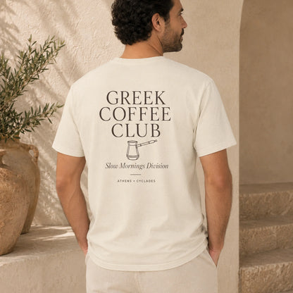 A man viewed from behind wearing a cream-colored t-shirt with a black graphic print that reads 'GREEK COFFEE CLUB' and 'Slow Mornings Division'. The setting is a warm, sunlit stone patio with a potted plant in the background.