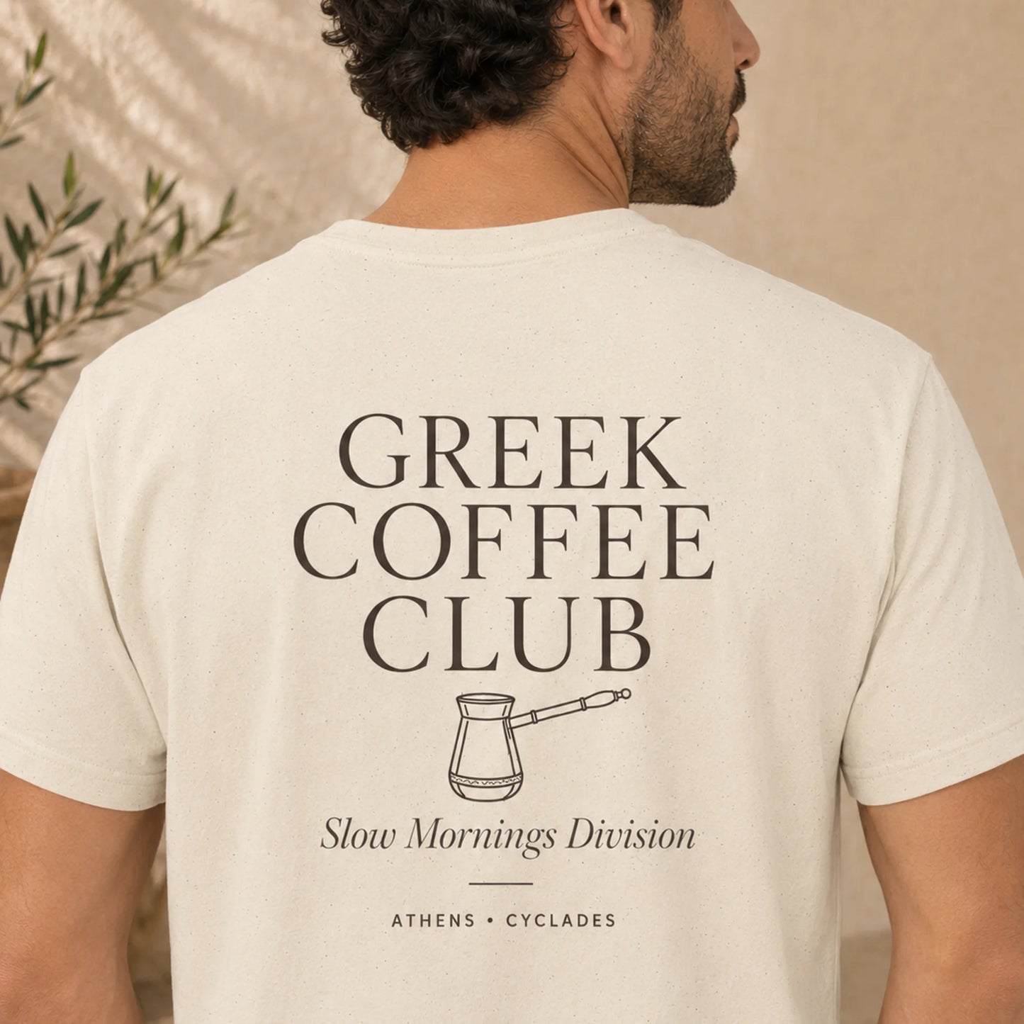 TP HERO: A man shown from the back wearing a cream-colored t-shirt with a black graphic print that reads 'GREEK COFFEE CLUB' and 'Slow Mornings Division'.