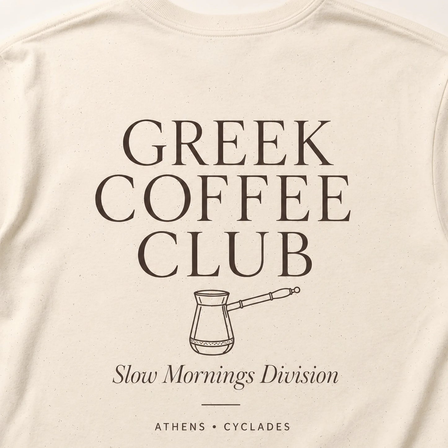 Close-up of an off-white or cream-colored t-shirt featuring a dark brown graphic print that reads 'GREEK COFFEE CLUB' with a coffee pot illustration and 'Slow Mornings Division' underneath.