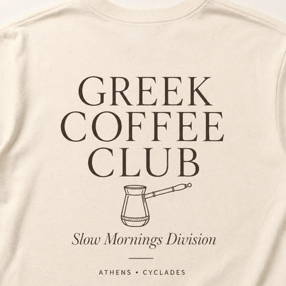 Close-up of an off-white or cream-colored t-shirt featuring a dark brown graphic print that reads 'GREEK COFFEE CLUB' with a coffee pot illustration and 'Slow Mornings Division' underneath.