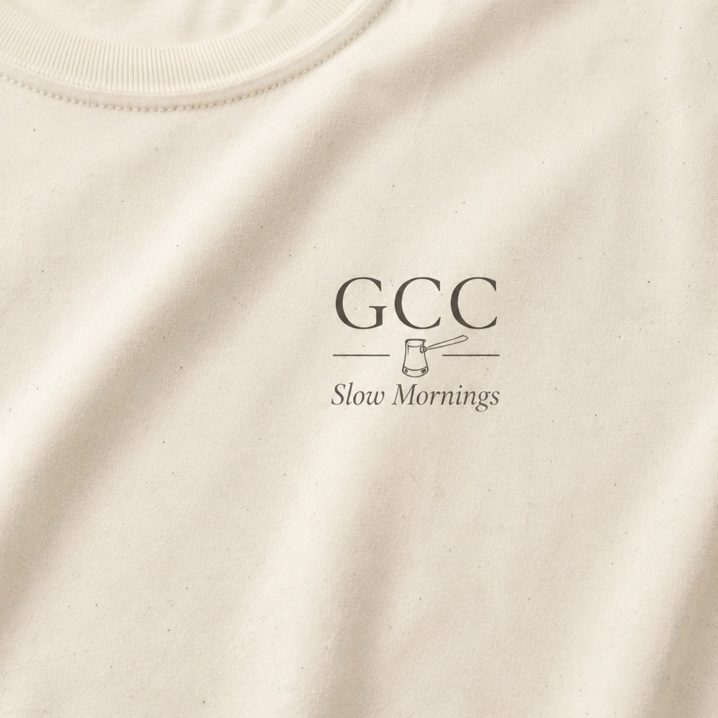 Close-up of an off-white crewneck t-shirt featuring a dark gray printed graphic on the chest with the text 'GCC' and 'Slow Mornings' alongside a small gavel illustration.