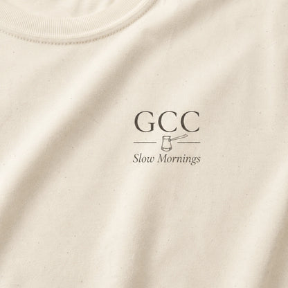 Close-up of an off-white crewneck t-shirt featuring a dark gray printed graphic on the chest with the text 'GCC' and 'Slow Mornings' alongside a small gavel illustration.