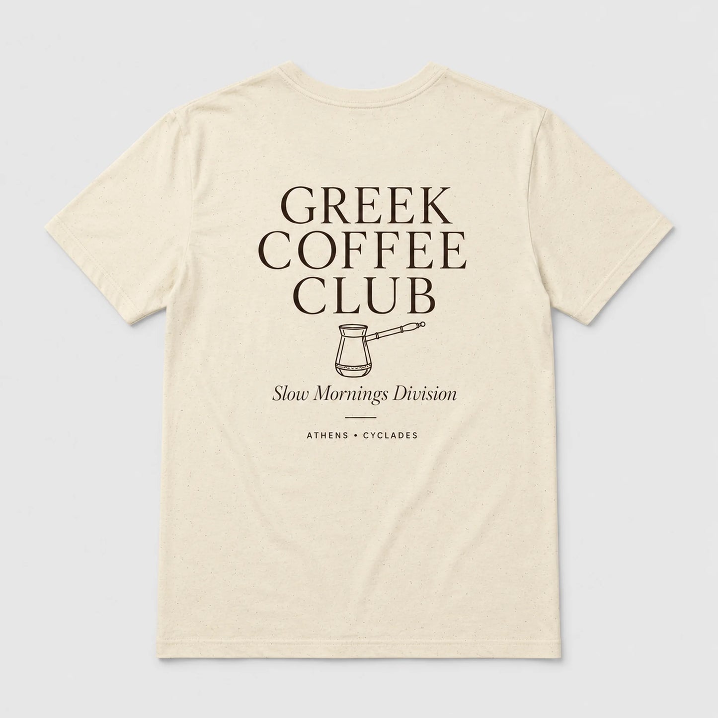 An off-white short-sleeve t-shirt with a dark brown graphic print on the front reading 'GREEK COFFEE CLUB' with a coffee pot illustration and 'Slow Mornings Division' underneath, shown on a light gray background.
