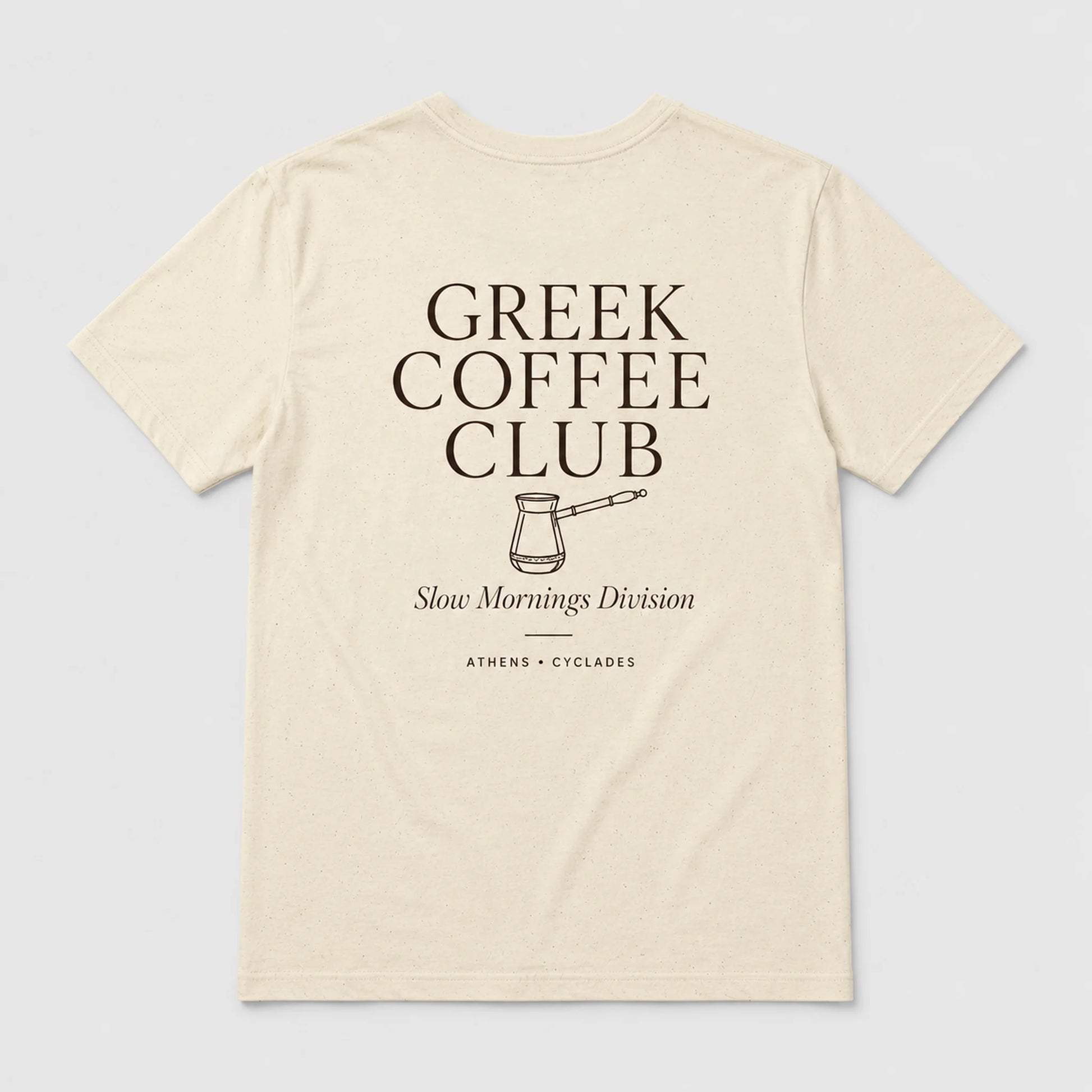 An off-white short-sleeve t-shirt with a dark brown graphic print on the front reading 'GREEK COFFEE CLUB' with a coffee pot illustration and 'Slow Mornings Division' underneath, shown on a light gray background.