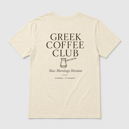 An off-white short-sleeve t-shirt with a dark brown graphic print on the front reading 'GREEK COFFEE CLUB' with a coffee pot illustration and 'Slow Mornings Division' underneath, shown on a light gray background.