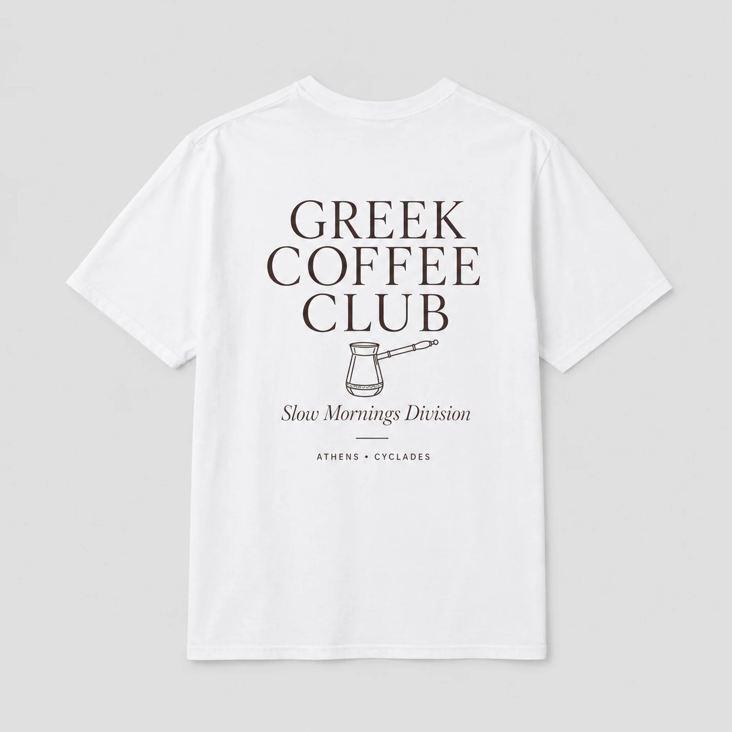 A white short-sleeve t-shirt with a dark brown graphic print on the front reading 'GREEK COFFEE CLUB' with a coffee pot illustration and 'Slow Mornings Division' underneath, shown on a light gray background.