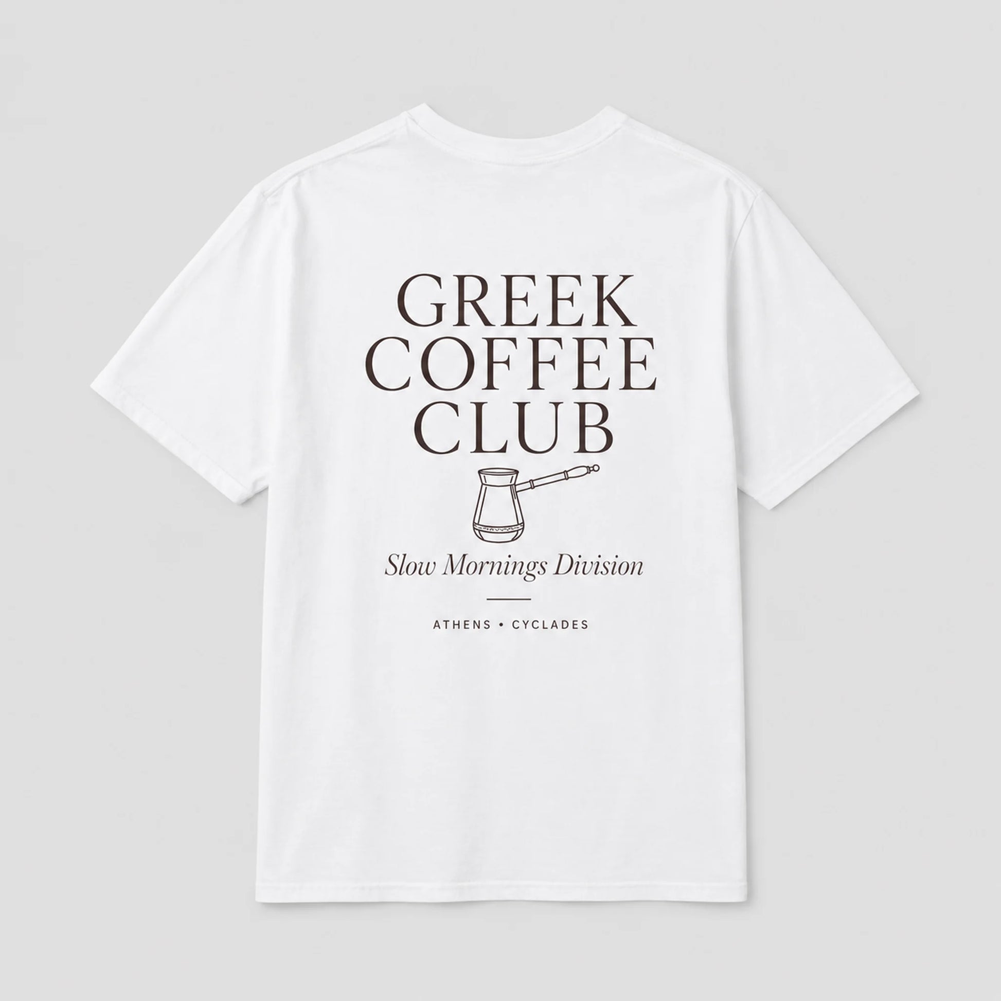 A white short-sleeve t-shirt with a dark brown graphic print on the front reading 'GREEK COFFEE CLUB' with a coffee pot illustration and 'Slow Mornings Division' underneath, shown on a light gray background.