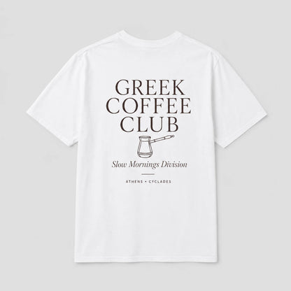 A white short-sleeve t-shirt with a dark brown graphic print on the front reading 'GREEK COFFEE CLUB' with a coffee pot illustration and 'Slow Mornings Division' underneath, shown on a light gray background.