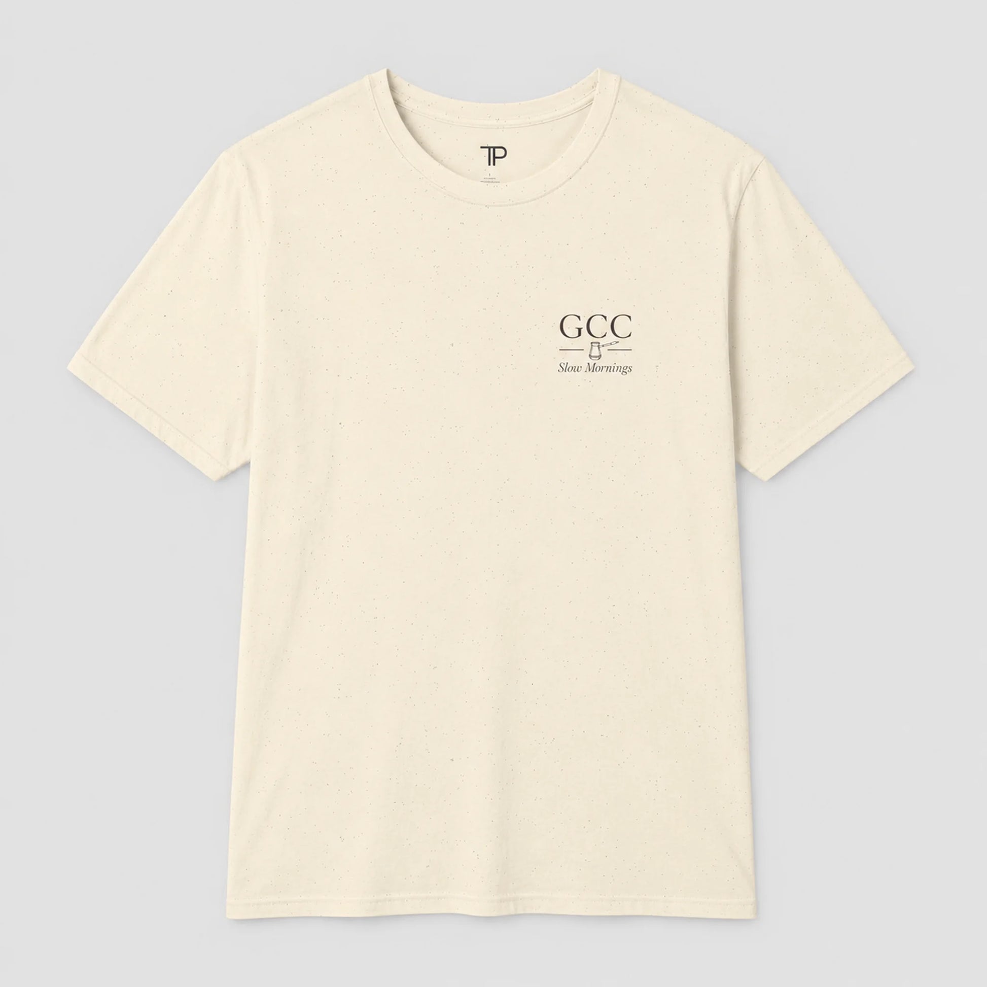 An off-white crewneck t-shirt with a small black graphic on the left chest reading 'GCC New Meersburg' and a small 'TP' logo inside the collar, displayed on a neutral gray background.