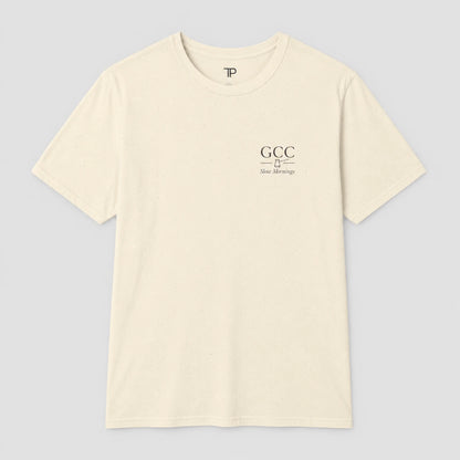 An off-white crewneck t-shirt with a small black graphic on the left chest reading 'GCC New Meersburg' and a small 'TP' logo inside the collar, displayed on a neutral gray background.