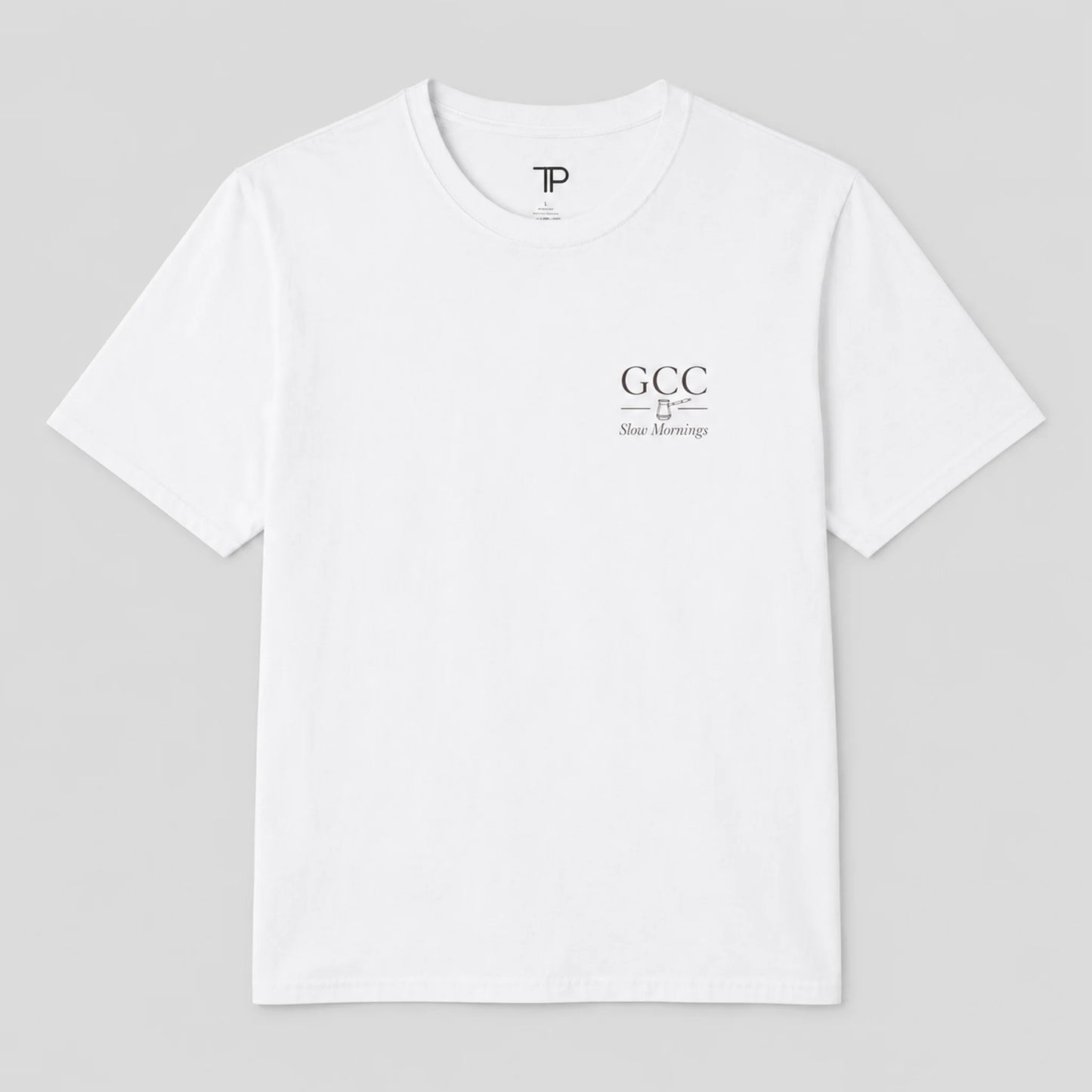 A white crewneck t-shirt with a small black graphic on the left chest reading 'GCC New Meersburg' and a small 'TP' logo inside the collar, displayed on a neutral gray 
