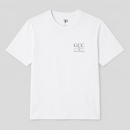 A white crewneck t-shirt with a small black graphic on the left chest reading 'GCC New Meersburg' and a small 'TP' logo inside the collar, displayed on a neutral gray 