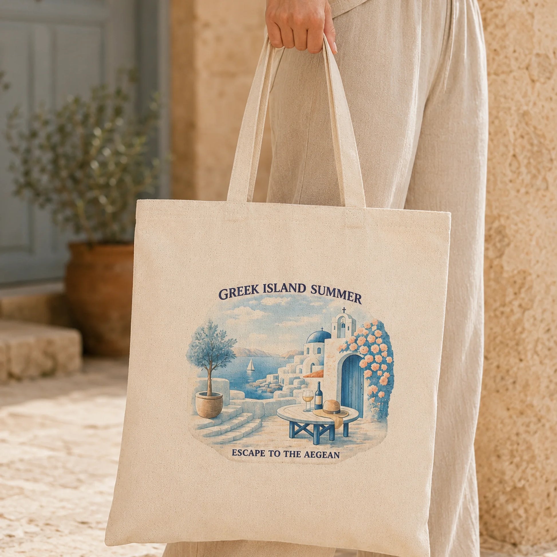 TP HERO: A person in a beige dress holds a cream canvas tote bag featuring a blue and white illustration of a Greek island summer scene with the text 'GREEK ISLAND SUMMER' and 'ESCAPE TO THE AEGEAN'.