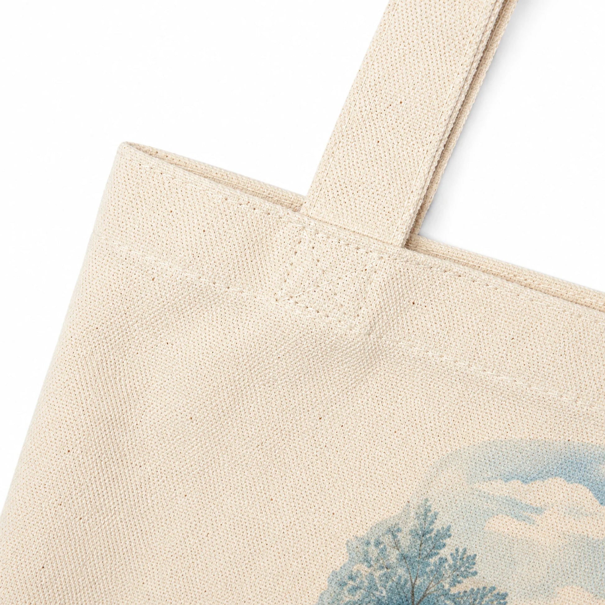 Close-up of a natural canvas tote bag showing the fabric texture, reinforced stitching, and a partial blue landscape print with a tree and sky.