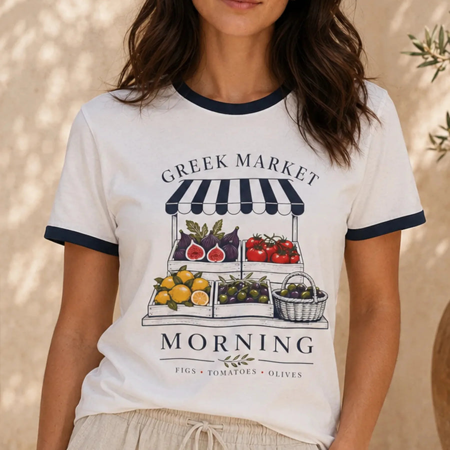 TP HERO: A woman wearing a white ringer t-shirt with navy blue trim and a graphic print of a Greek market morning produce stand.