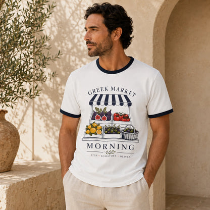 TP HERO: A man wearing a white ringer t-shirt with navy blue trim and a colorful 'Greek Market Morning' graphic print, standing in a sunlit Mediterranean-style stone corridor.