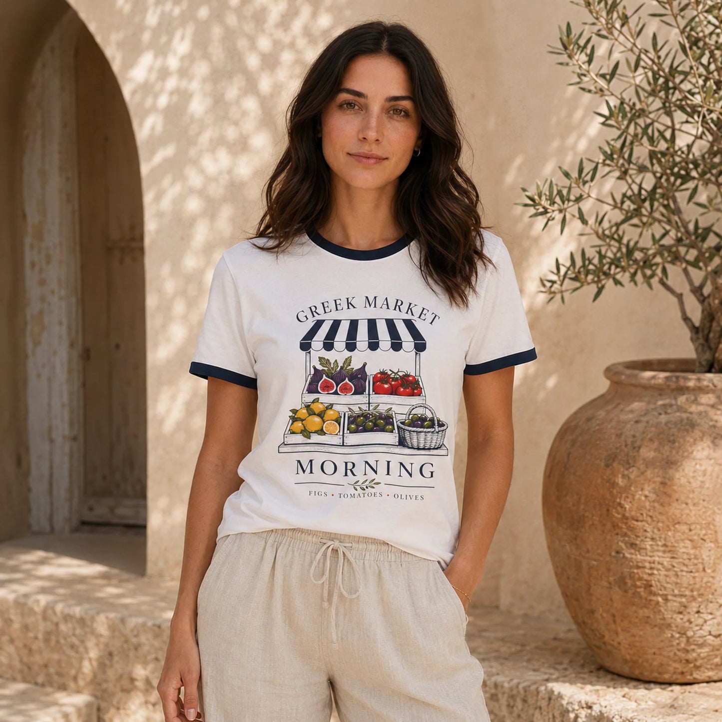 A woman stands outdoors wearing a white ringer T-shirt with navy trim and a colorful Greek market graphic on the front, paired with light beige drawstring shorts. The background features a warm beige stone wall, an archway, and a large terracotta planter with olive branches.