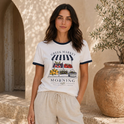 A woman stands outdoors wearing a white ringer T-shirt with navy trim and a colorful Greek market graphic on the front, paired with light beige drawstring shorts. The background features a warm beige stone wall, an archway, and a large terracotta planter with olive branches.