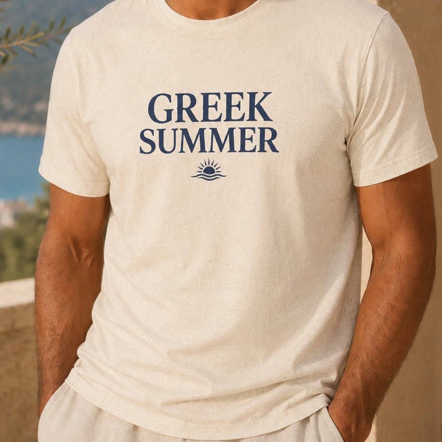 A man wearing a cream-colored t-shirt with 'GREEK SUMMER' printed in navy blue text and a small sun-and-wave graphic below it, set against a blurred outdoor background.