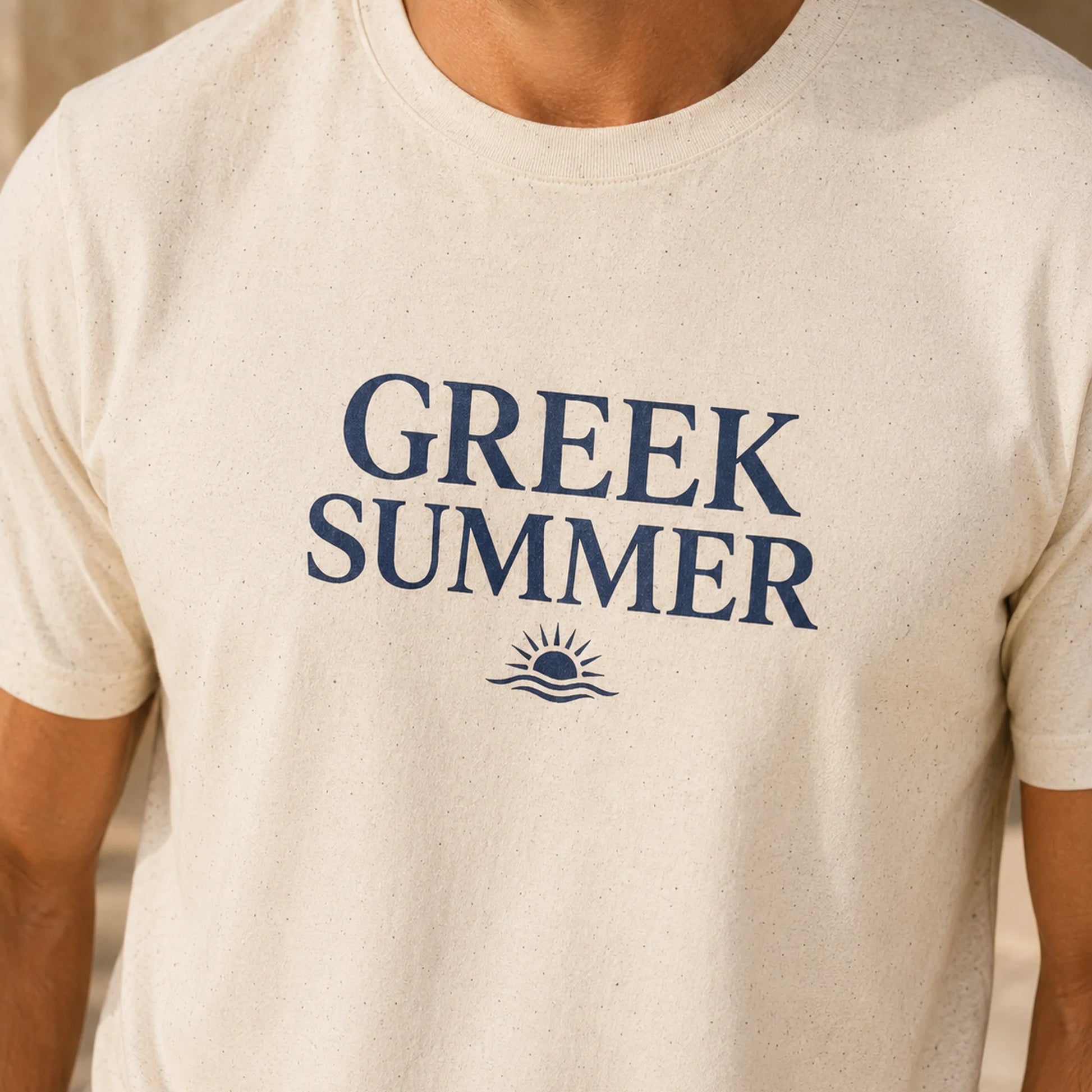 A close-up of a person wearing a light beige speckled t-shirt with 'GREEK SUMMER' printed in navy blue text and a small sun graphic on the chest.