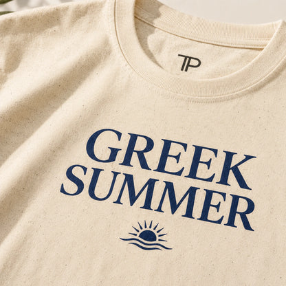 Close-up of a cream-colored textured t-shirt featuring the words 'GREEK SUMMER' in navy blue text with a small sun and wave graphic underneath. The inside collar shows a 'TP' logo.