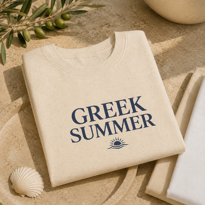 A folded cream-colored cotton t-shirt with 'GREEK SUMMER' printed in navy blue text and a small sun-and-wave graphic, styled on a beige stone surface with olive branches, a seashell, and a folded white towel.