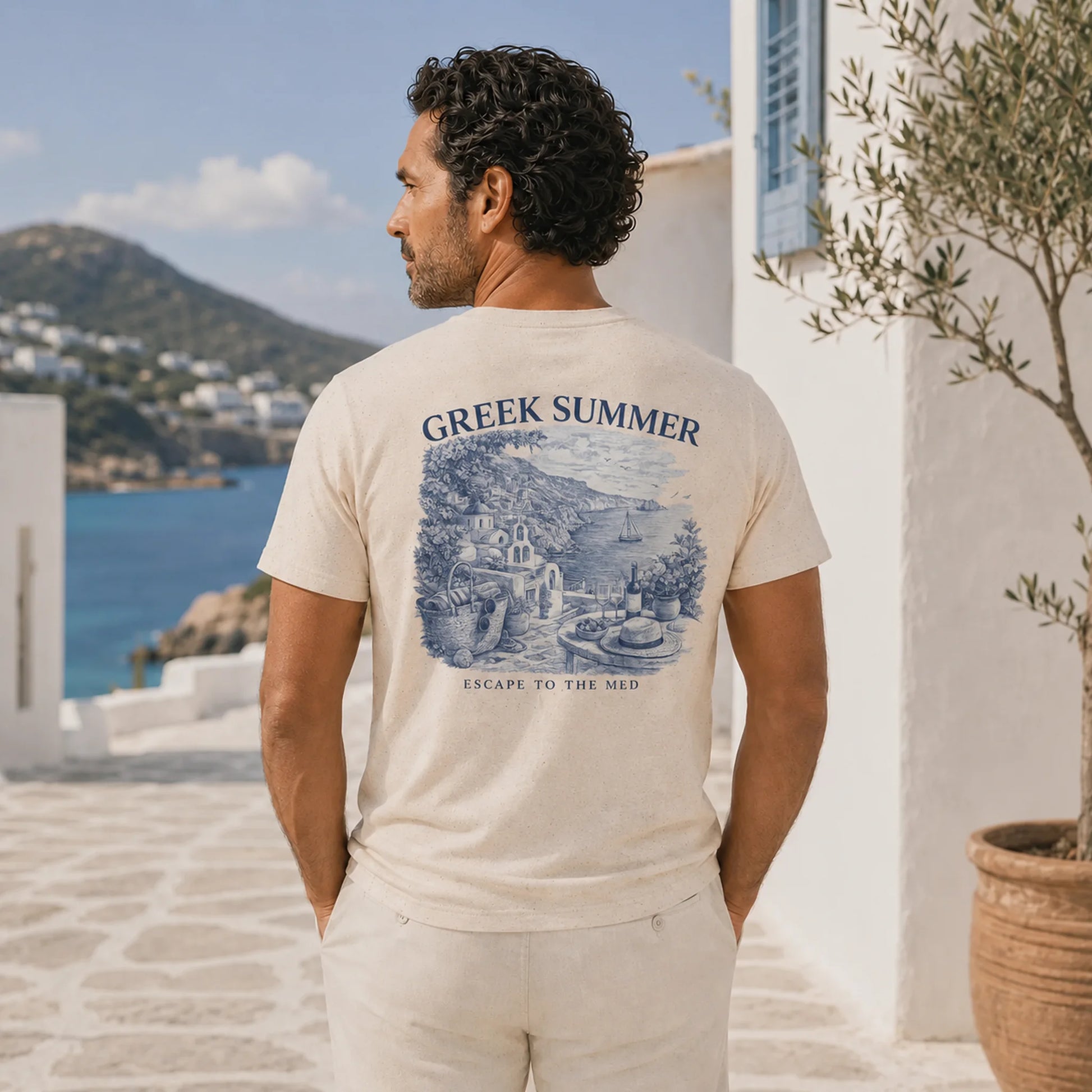 Man wearing a 'Greek Summer' t-shirt with a scenic design, standing in a picturesque outdoor setting.