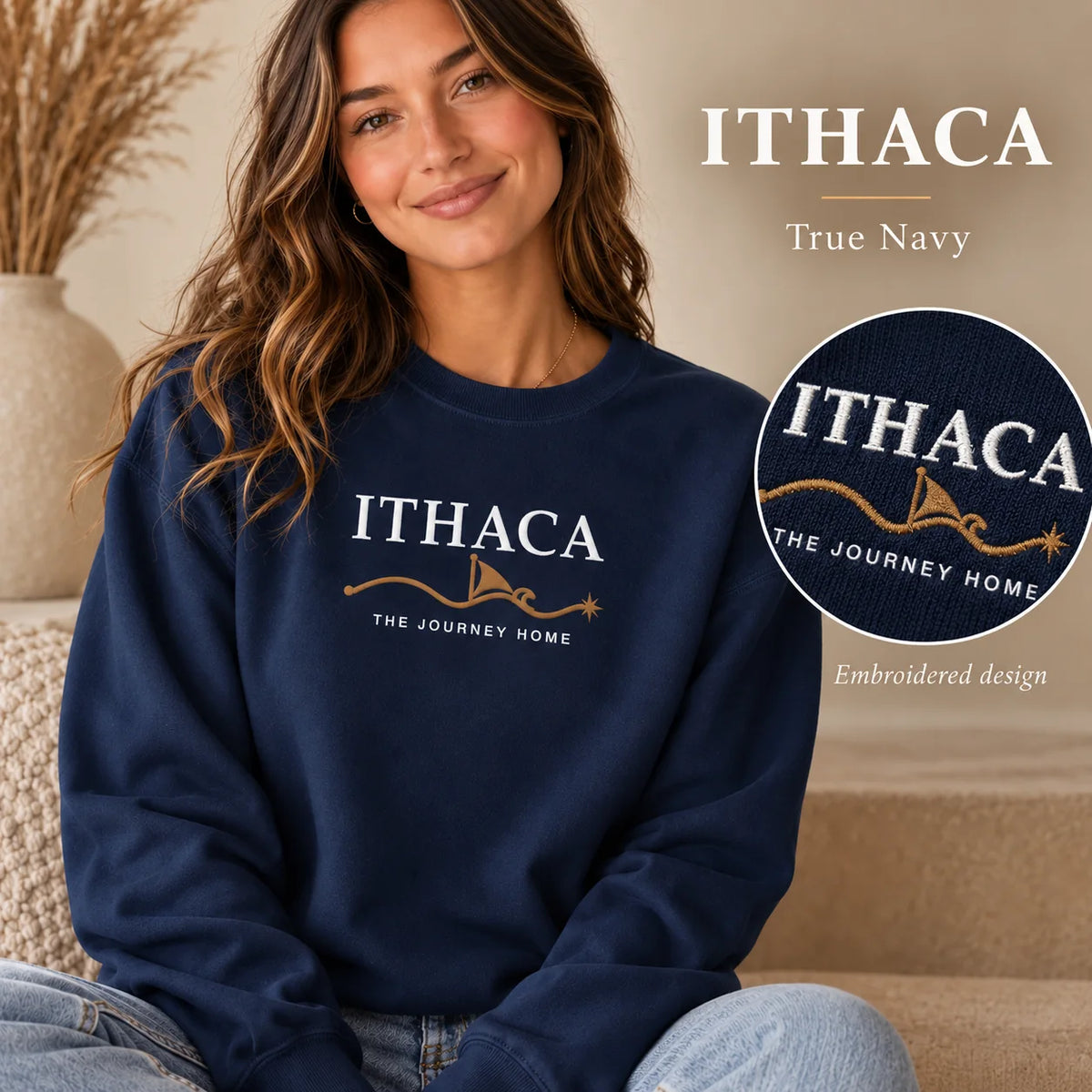 A woman wearing a navy crewneck sweatshirt with an embroidered Ithaca 'The Journey Home' design, shown in a cozy indoor setting with text overlays highlighting the true navy color and the embroidered detail.