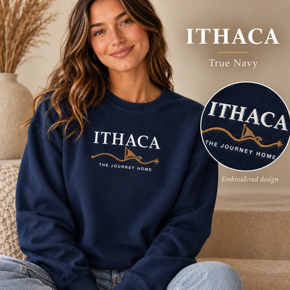 A woman wearing a navy crewneck sweatshirt with an embroidered Ithaca 'The Journey Home' design, shown in a cozy indoor setting with text overlays highlighting the true navy color and the embroidered detail.