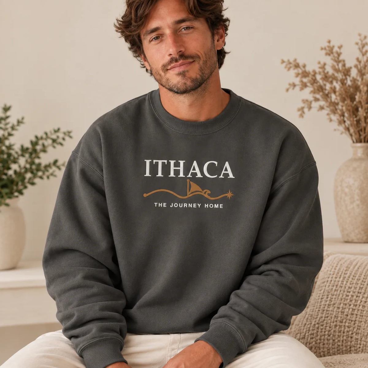 A man wearing a dark gray crewneck sweatshirt with 'ITHACA THE JOURNEY HOME' embroidered in white and gold on the front, sitting in a cozy indoor setting.