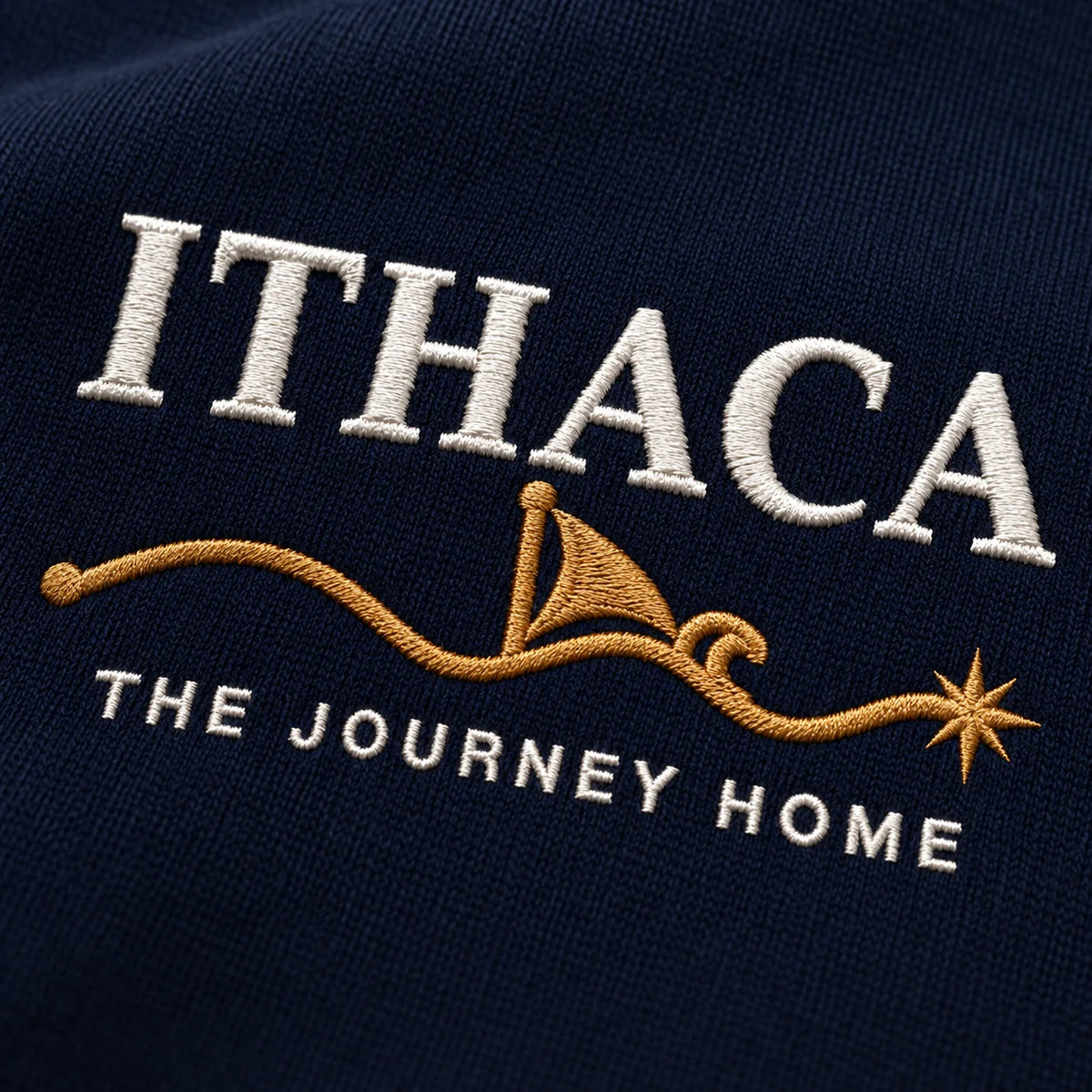 Close-up of embroidered text reading 'ITHACA' in white, a gold sailboat and wave motif, and 'THE JOURNEY HOME' in white on a dark navy fabric background.
