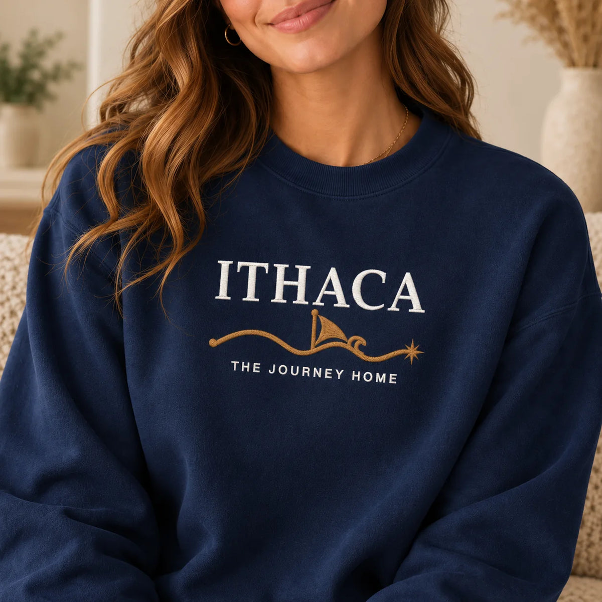 TP HERO: A woman wearing a navy blue crewneck sweatshirt with a white and gold graphic that reads 'ITHACA THE JOURNEY HOME' on the chest, shown in a cozy indoor setting.