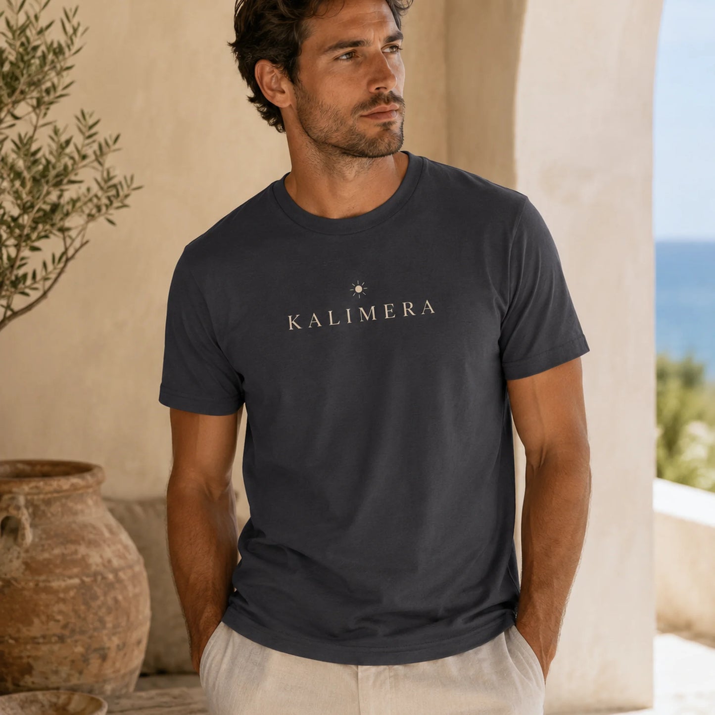 A man wearing a dark charcoal gray t-shirt with 'KALIMERA' printed in white across the chest, standing in a sunlit outdoor setting with beige walls and a rustic pot.