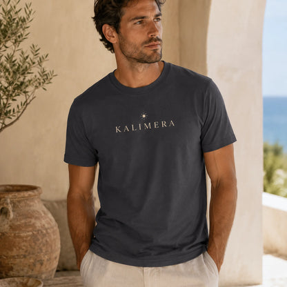 A man wearing a dark charcoal gray t-shirt with 'KALIMERA' printed in white across the chest, standing in a sunlit outdoor setting with beige walls and a rustic pot.