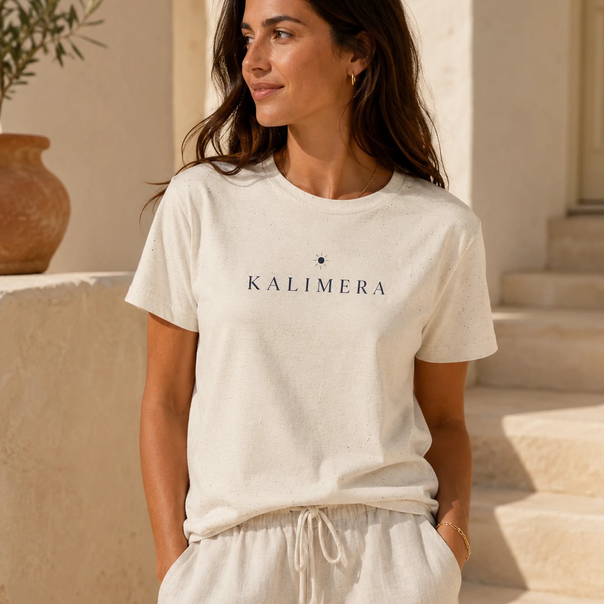 A woman wearing a natural-colored short-sleeve t-shirt with 'KALIMERA' printed in navy blue across the chest, standing in a warm, sunlit outdoor setting with beige stone steps and a terracotta pot in the background.