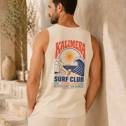 A man wearing a cream-colored tank top with a colorful 'KALIMERA SURF CLUB' graphic on the back, leaning against a textured beige wall in a sunlit outdoor setting.