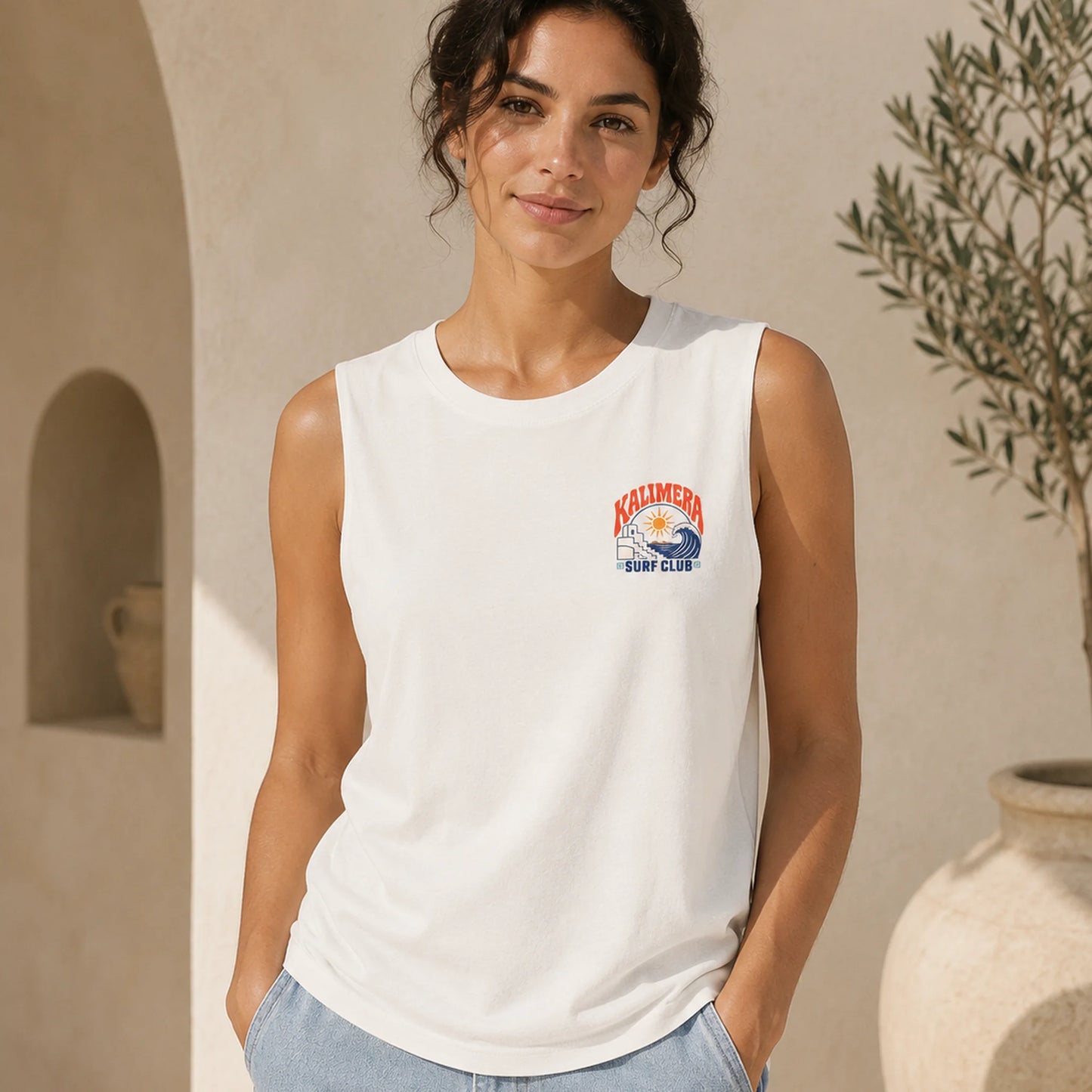 A woman wearing a white sleeveless tank top with a small colorful 'Kolbimer Surf Club' graphic on the left chest, posed in a warm, sunlit architectural setting with beige walls and a potted plant.