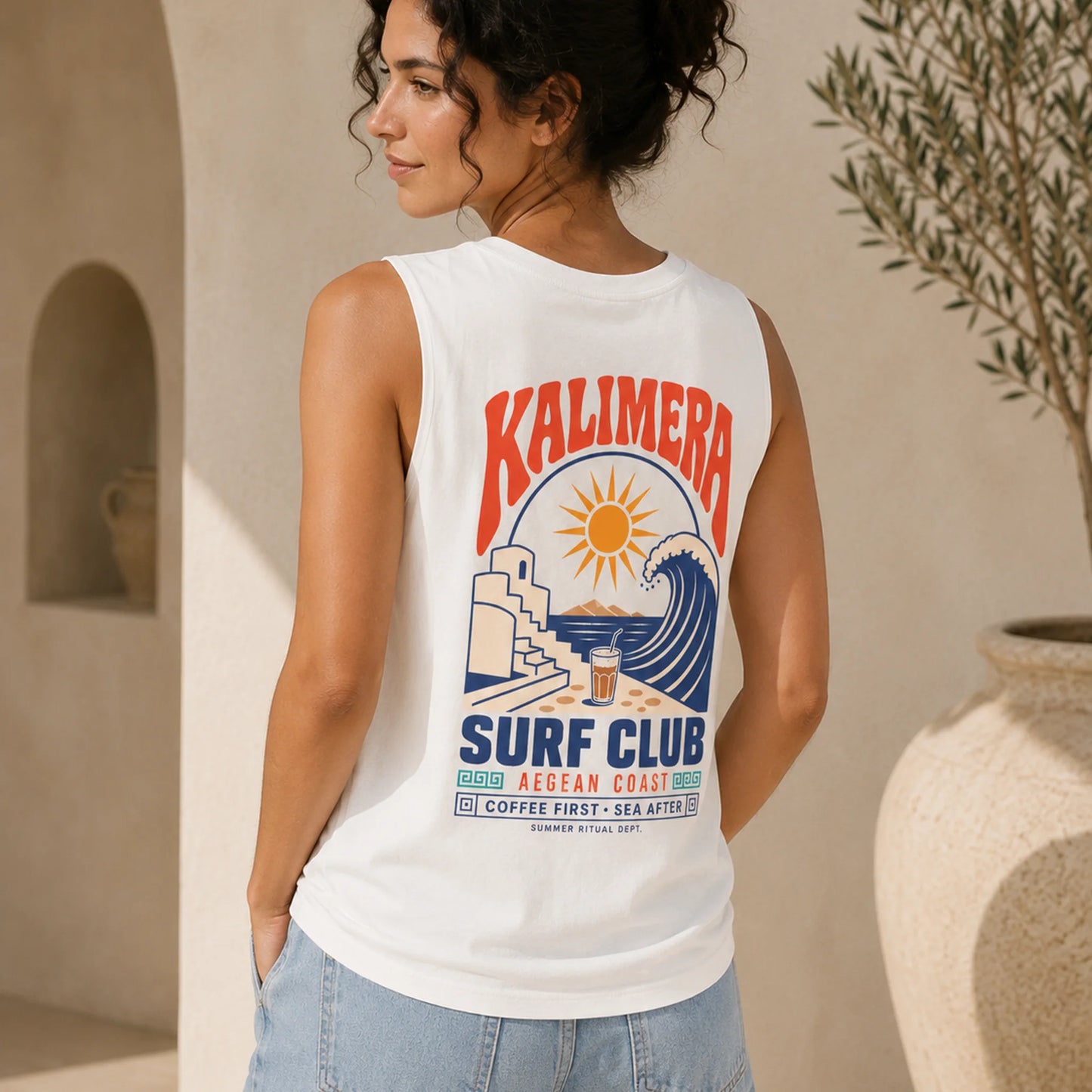 A woman wearing a white sleeveless tank top with a colorful retro-style 'KALIMERA SURF CLUB' graphic on the back, standing in a sunlit beige architectural setting.