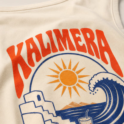 Close-up of a cream-colored t-shirt featuring a colorful graphic print with the word 'KALIMERA' in bold orange-red letters, a yellow sun, a blue wave, and a white building.