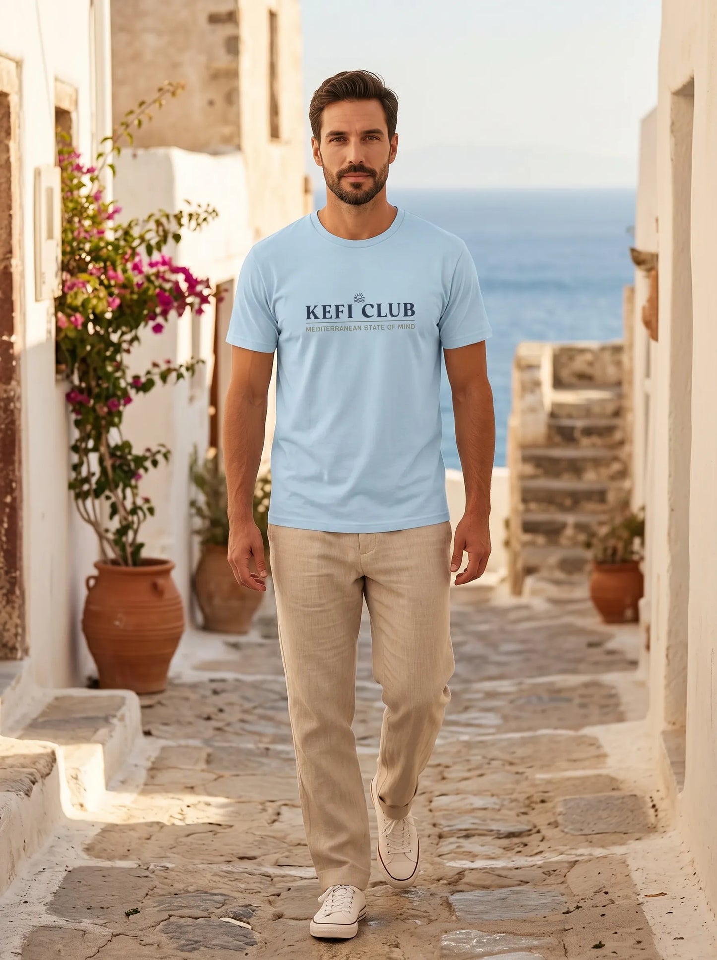 Man wearing a light blue t-shirt with 'KEFI CLUB' text, walking on a stone path with ocean view.