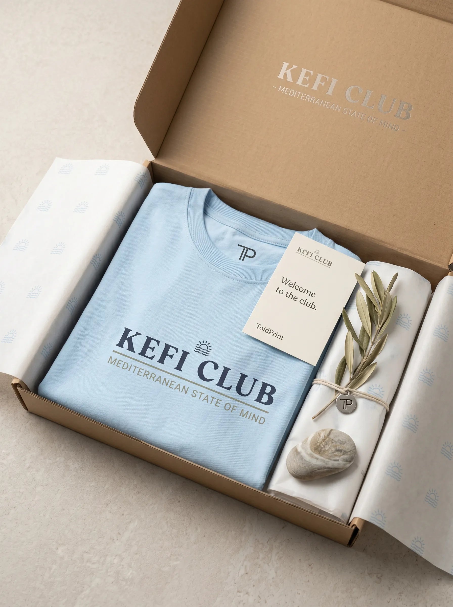 Open box containing a light blue t-shirt with 'Kefi Club' branding, a small plant, and a tag.