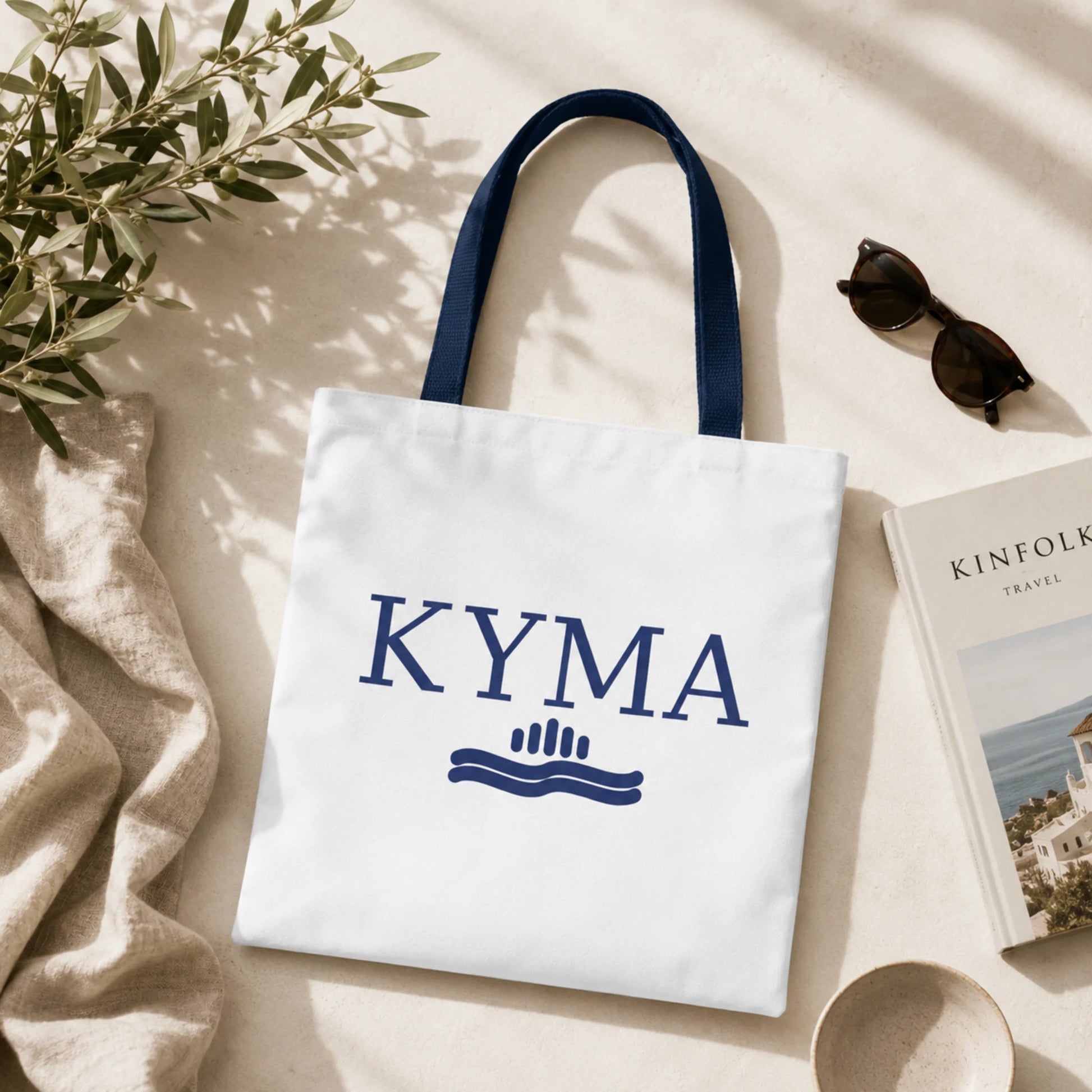 White tote bag with 'KYMA' branding on a light surface with a plant and sunglasses.