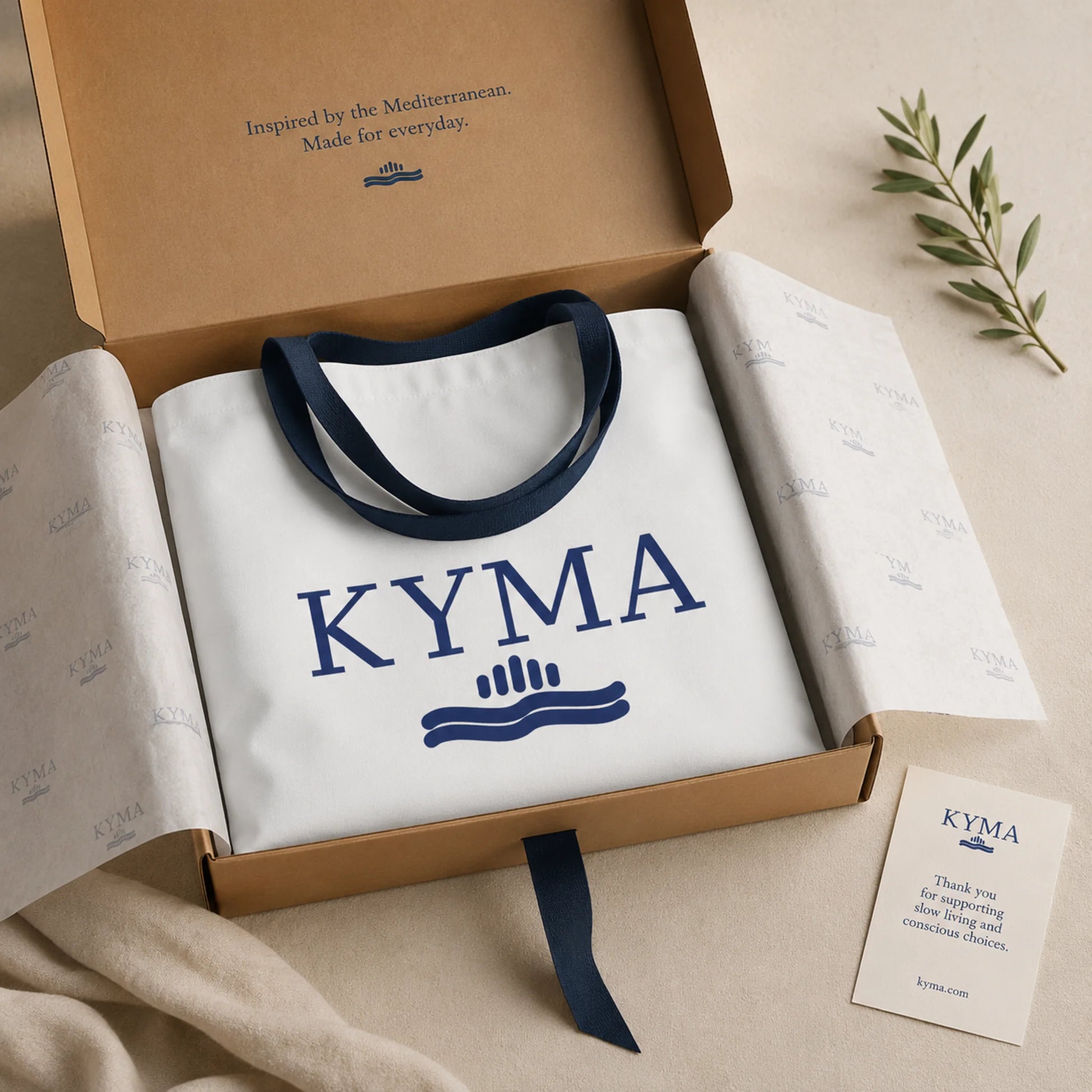 White tote bag with 'KYMA' branding in a box, accompanied by a card and greenery.
