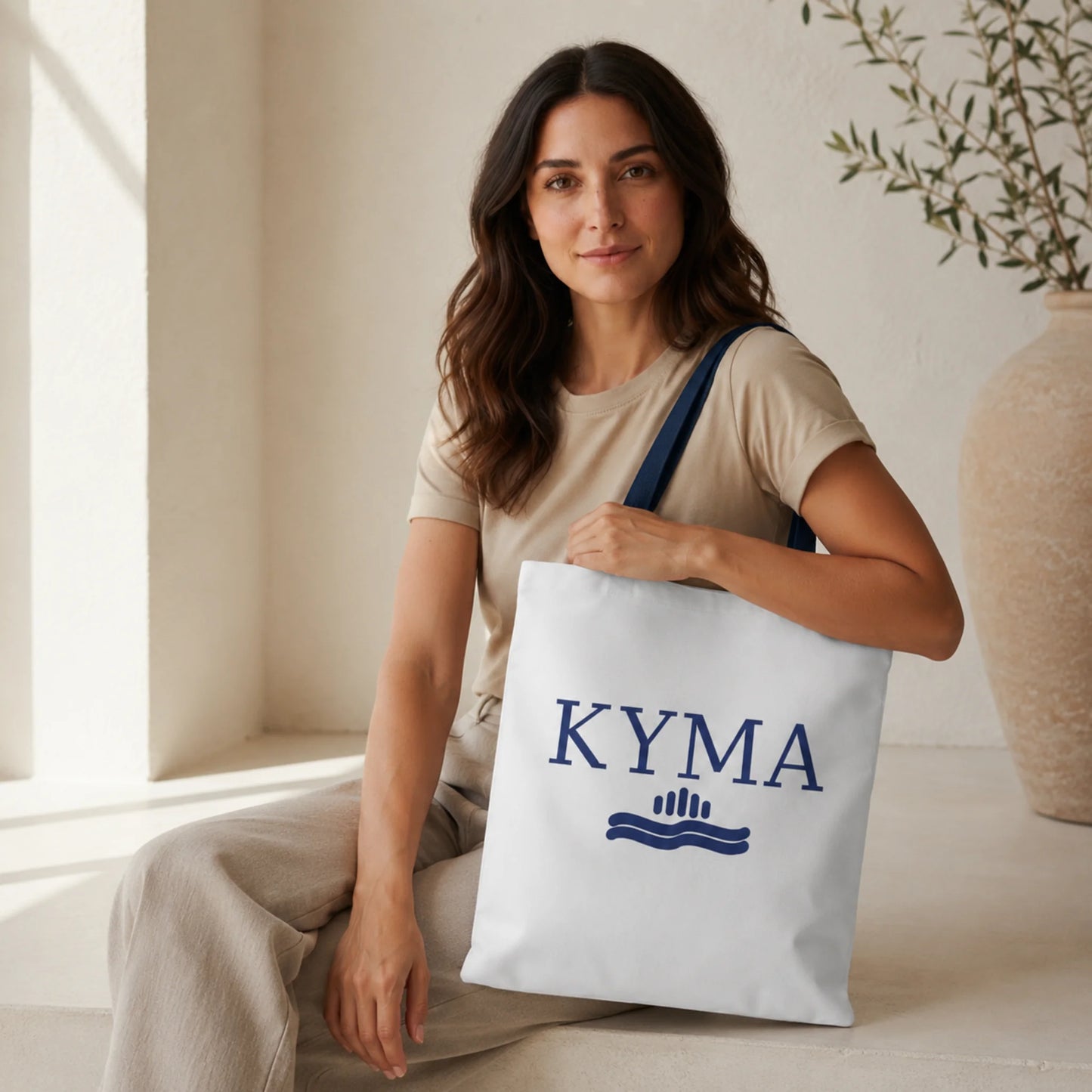 Woman holding a white tote bag with 'KYMA' branding in a home setting.