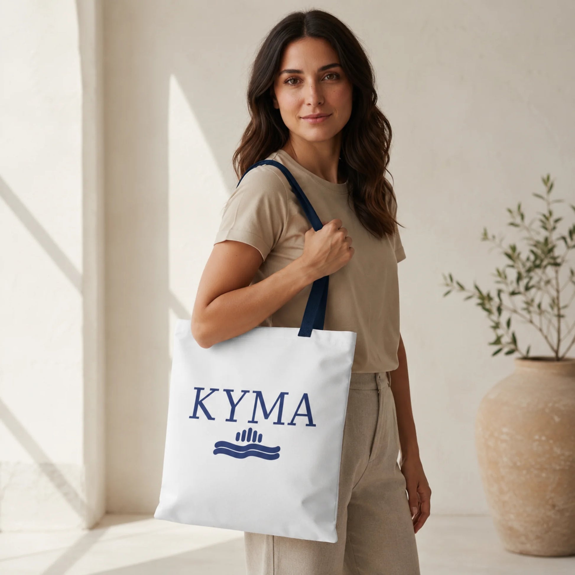 Woman holding a white tote bag with 'KYMA' branding in a bright indoor setting.