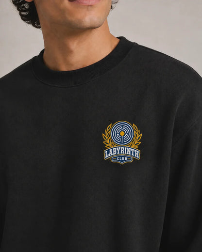 A close-up of a person wearing a black crewneck sweatshirt with a blue and gold embroidered 'Labyrinth Club' logo on the left chest, set against a neutral gray background.