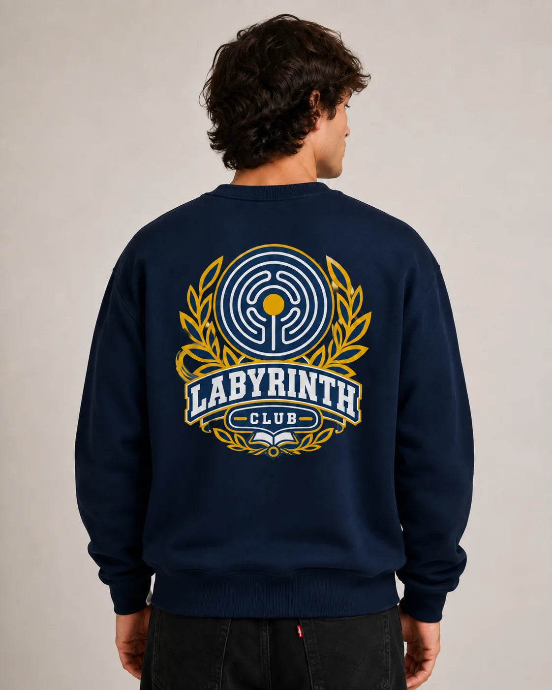 A man viewed from behind wearing a navy blue crewneck sweatshirt with a large yellow and white 'LABYRINTH CLUB' graphic on the back, paired with black jeans against a neutral beige background.