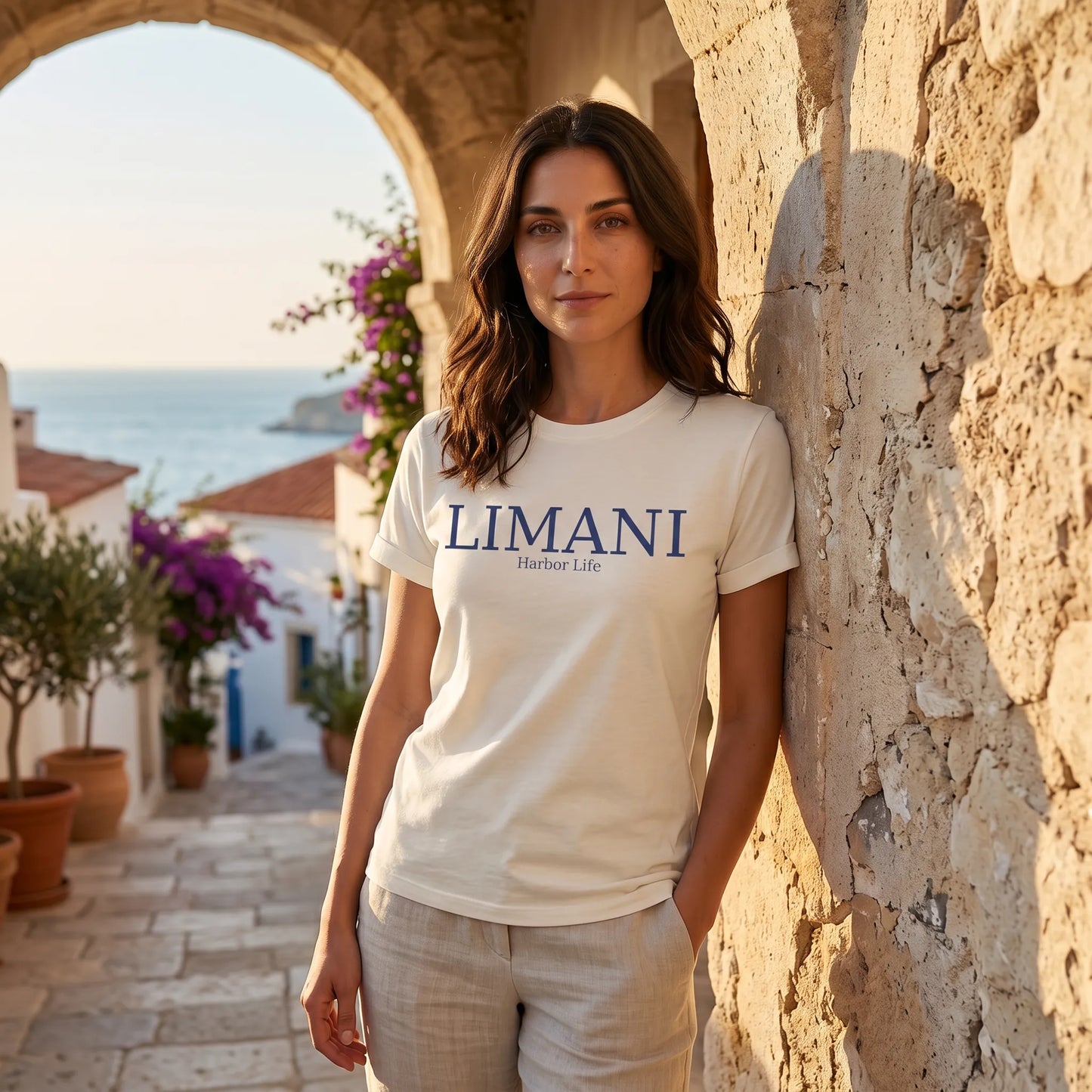Woman wearing a 'LIMANI' t-shirt standing against a stone wall with a scenic background.